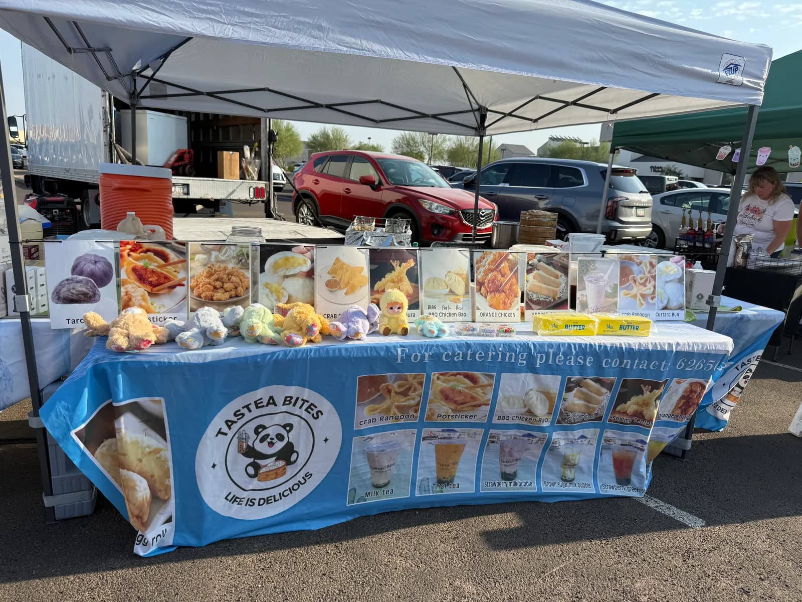The Tastea Bites stall on a market morning: sky-blue tablecloth, laminated menu cards clipped up, plush pandas on the counter
